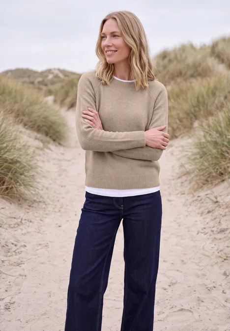 Eine Frau in beige Pullover und blauen Jeans steht mit verschränkten Armen lächelnd in Dünen mit Strandgras.