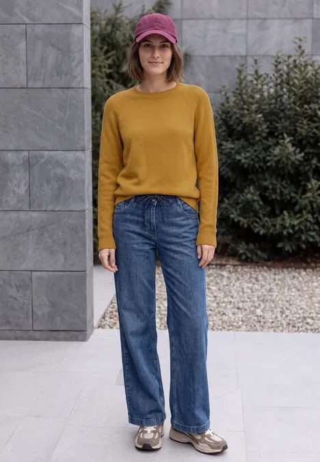 Eine Frau mit senfgelbem Pullover, blauen Jeans, Cap und Turnschuhen steht vor einem modernen Gebäude.
