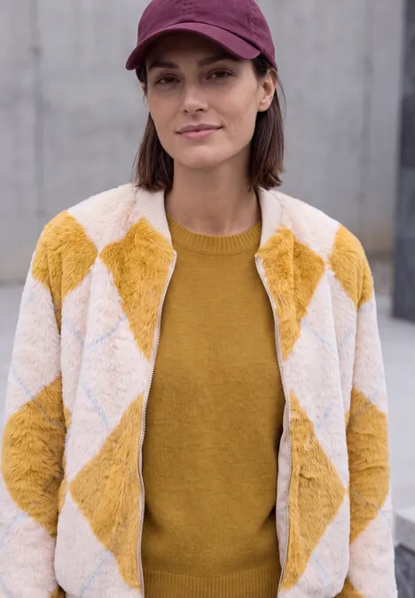 Eine Frau trägt eine gelb-weiße Rautenmusterjacke, einen Senfpullover und ein rotes Cap.