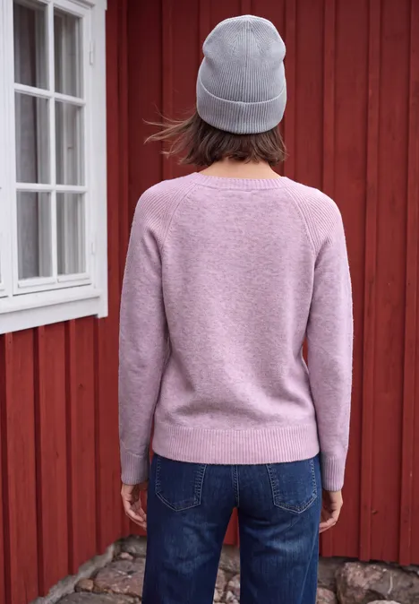Persoon met roze trui, grijze muts en spijkerbroek, rug naar kijker voor een rode houten wand.