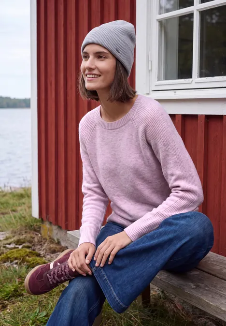 Vrouw lachtend in roze trui, spijkerbroek en grijze muts zittend op houten traptrede voor rood huis aan het meer.
