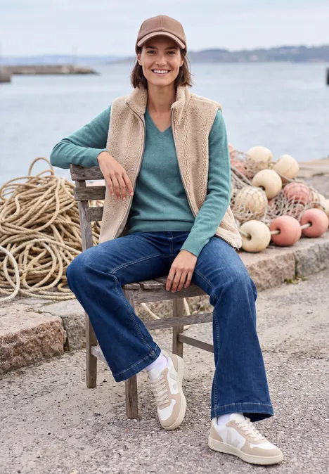 Vrolijke vrouw met pet, fleecevest en jeans zit bij de haven tussen touwen en drijvers.