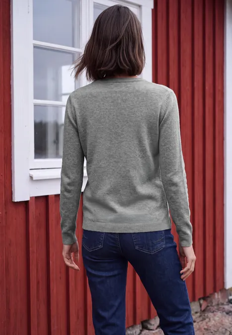 Een vrouw in een grijze trui en jeans, met haar rug naar de camera, voor een rode houten gevel.
