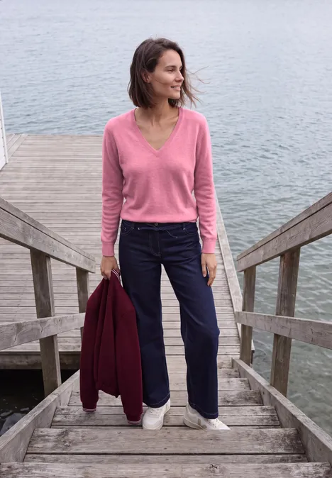 Een vrouw in een roze trui en marineblauw broek staat op houten treden bij het water met een rode jas.