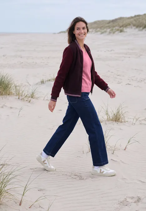 Vrouw in rode jas en spijkerbroek wandelt glimlachend over duinen op het strand.