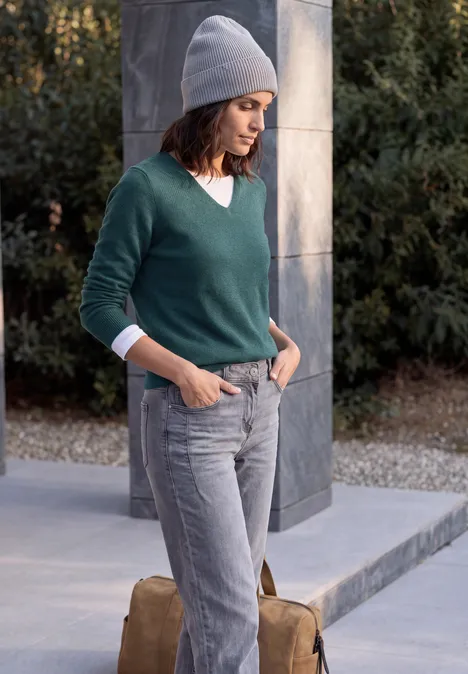Langarm Pullover mit V-Neck dark sea green melange Frau in grauer Mütze, grünem Pullover und grauen Jeans lehnt an Säule im Freien.