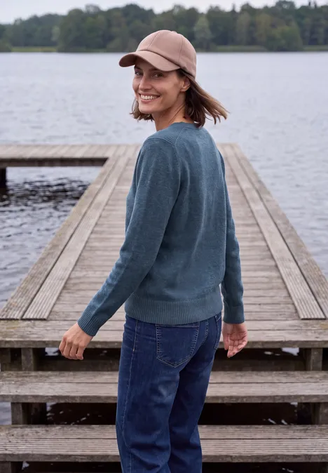 Langarm Pullover mit V-Neck cotton blue melange Eine lächelnde Frau mit Mütze und Pullover steht auf einem Steg am ruhigen See.