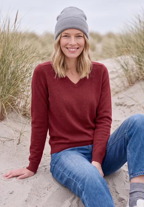 Een glimlachende vrouw met muts, bordeaux trui en spijkerbroek zittend in het zand bij duinen.