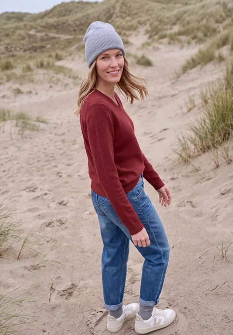 Jonge vrouw in rode trui, spijkerbroek en grijze muts glimlacht in de duinen.