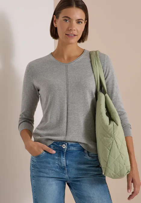 Eine Frau trägt einen grauen Strickpullover, blaue Jeans und eine olivgrüne Stepptasche über der Schulter.