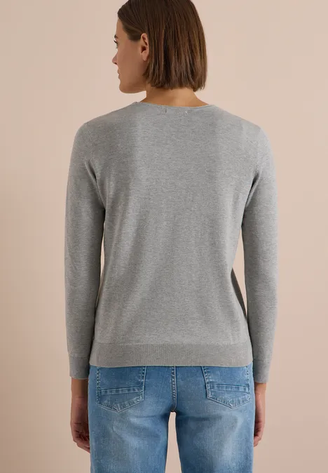 Frau von hinten in grauem Pullover und blauen Jeans vor beigem Hintergrund.