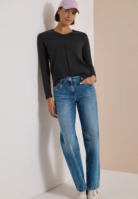 Frau lehnt an Wand in schwarzem Pullover, blauen Jeans und rosa Cap.