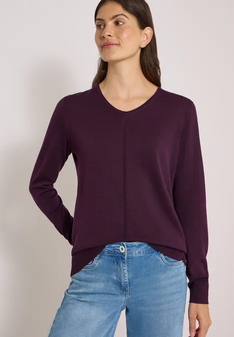 Eine Frau trägt einen dunkelvioletten V-Ausschnitt-Pullover und blaue Jeans vor hellem Hintergrund.