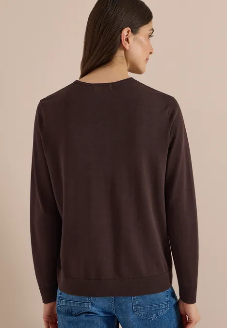 Frau von hinten in braunem Pullover und blauer Jeans vor neutralem Hintergrund.