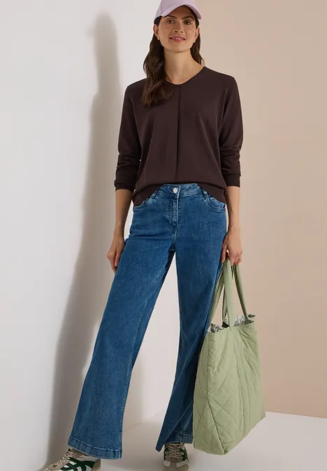 Frau in kastanienbraunem Langarmshirt, weiter Jeans mit vorderen Schlitzen, weißen Sneakern und großer grüner Tasche.
