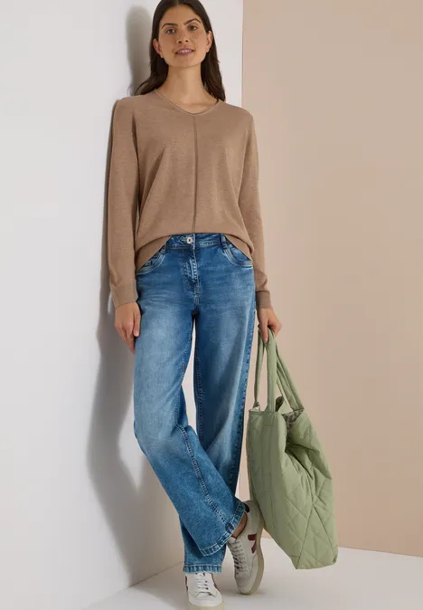 Frau in beigem Pullover, blauen Jeans und weißen Sneakers hält grüne Stepp-Tasche vor neutralem Hintergrund.