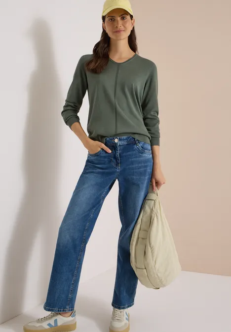 Frau in grünem Pullover, blauen Jeans, weißen Sneakern und gelber Mütze, mit leichter Tasche.