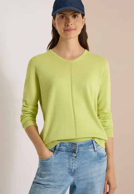 Frau in hellgrünem Pullover, Jeans und Baseballcap vor neutralem Hintergrund.
