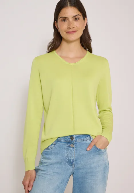 Frau in hellgrünem Pullover und blauen Jeans, Hand in Tasche, lächelnd vor neutralem Hintergrund.