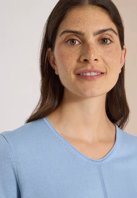 Porträt einer lächelnden Frau mit dunklem Haar in einem hellblauen Shirt vor hellem Hintergrund.
