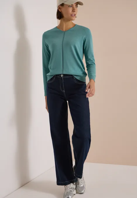 Eine Frau in grüner Strickjacke, Jeans und Sneakern vor hellem Hintergrund.