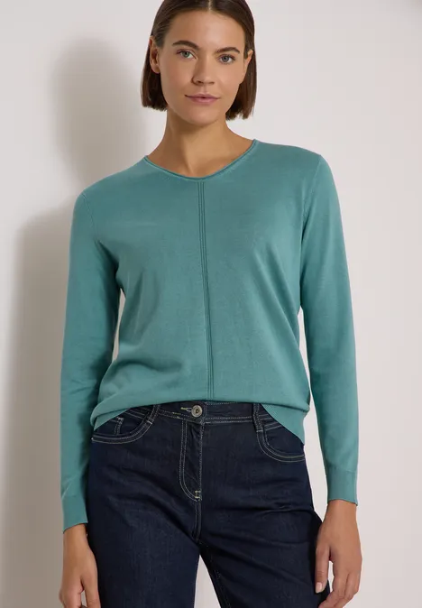 Frau trägt tealfarbenen Pullover und dunkle Jeans, steht vor hellem Hintergrund.