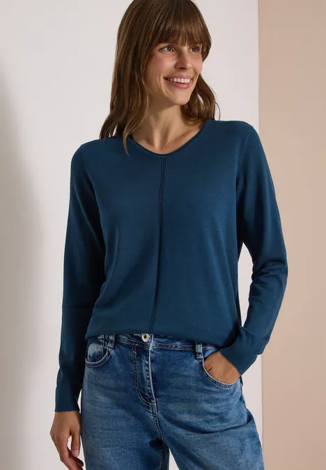 Basic Pullover mit V-Neck in Unifarbe moonlight blue Frau mit blauem Pullover und Jeans steht lächelnd vor hellem Hintergrund.