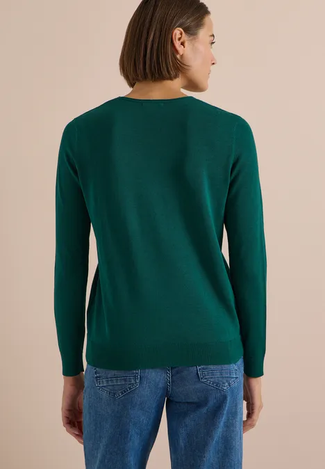 Rückansicht einer Frau in grünem Pullover und blauen Jeans vor beigem Hintergrund.