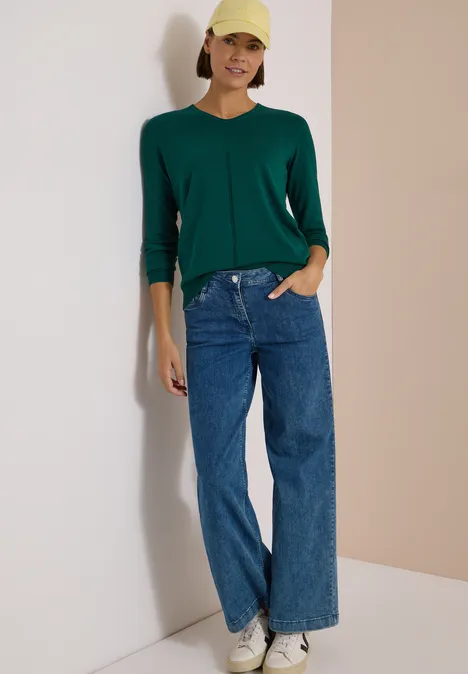 Eine Frau lehnt lässig an einer Wand, trägt einen grünen Cardigan, weite Jeans und eine gelbe Mütze.