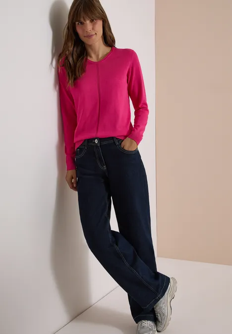 Basic Pullover mit V-Neck in Unifarbe pink flash Frau lehnt an Wand, trägt pinken Pullover, weite Jeans und weiße Sneaker vor neutralem Hintergrund.