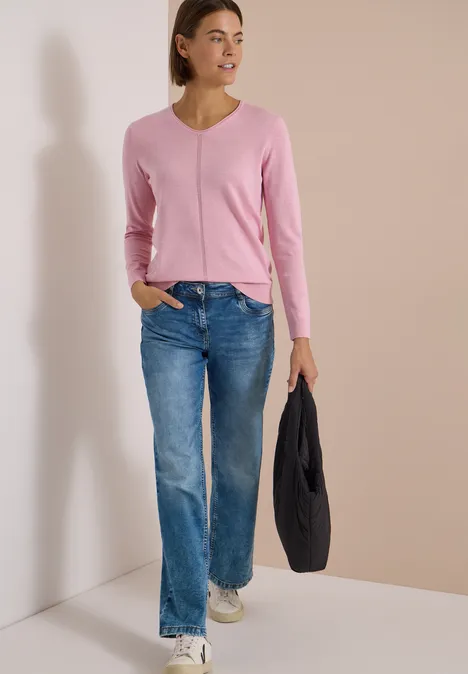 Frau in pinkem V-Pullover, Jeans und Sneakern, trägt schwarze Jacke, steht vor neutralem Hintergrund.