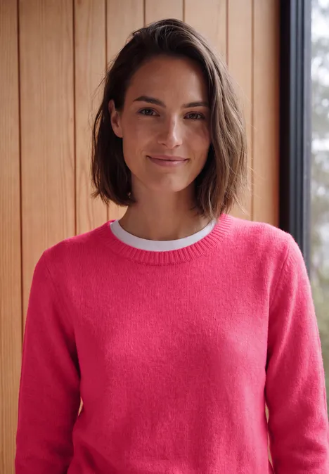Vrouw met bruin haar lacht en draagt een roze trui voor een houten wand.