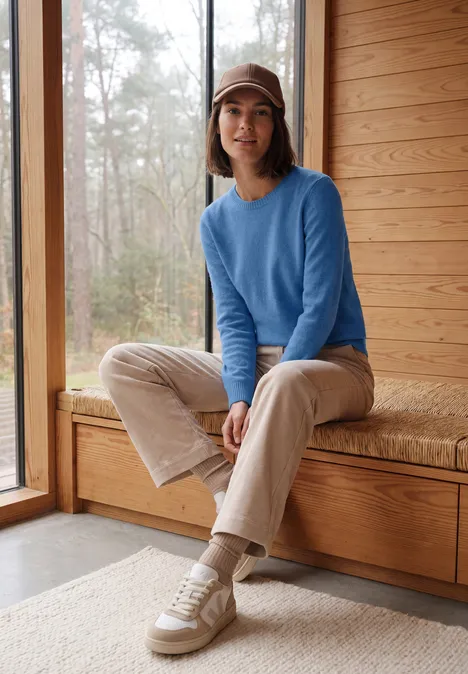 Vrouw in blauwe trui en pet zit op een houten bank in een lichte ruimte met uitzicht op het bos.