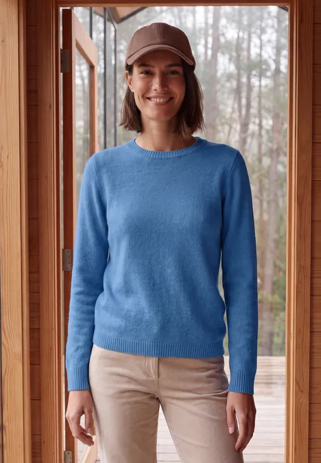 Lachende vrouw in blauw truitje, beige broek en pet, staand voor een houten raam.