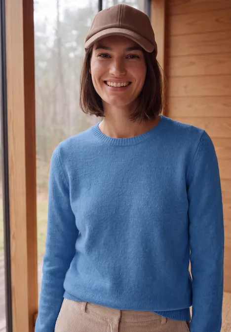 Jonge vrouw met blauwe trui en beige pet voor een houten wand, ze lacht.