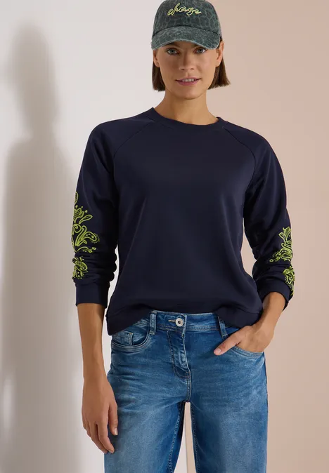 Frau in navyblauem Sweatshirt mit neongrünen Blumenstickereien an den Ärmeln, Jeans und Basecap.