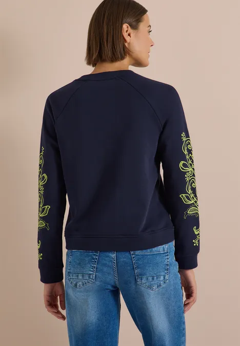 Frau von hinten in dunkelblauem Sweatshirt mit grünen Ärmeldetails und blauen Jeans.