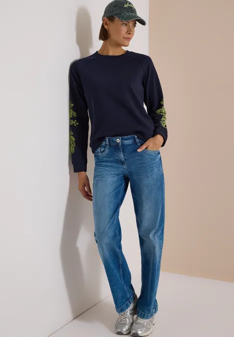 Frau in dunkelblauem Pullover mit grünen Ärmeldetails, blauen Jeans, Cap und Sneakern.