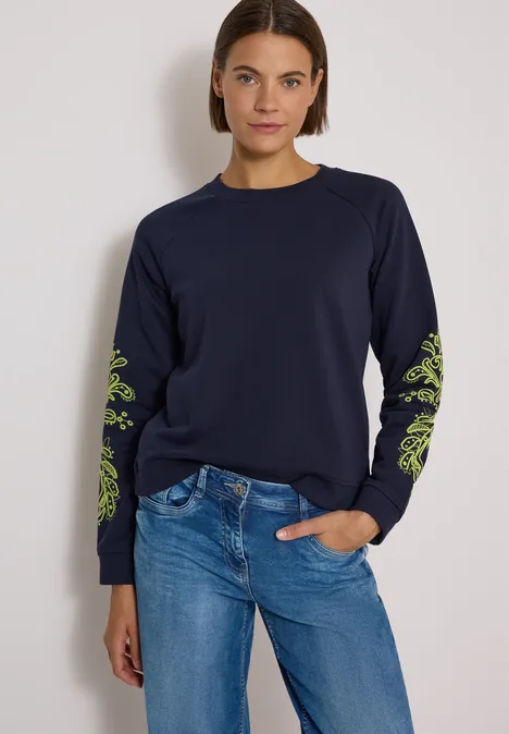 Frau in dunkelblauem Sweatshirt mit grünen Ärmelmustern und Jeans vor hellem Hintergrund.