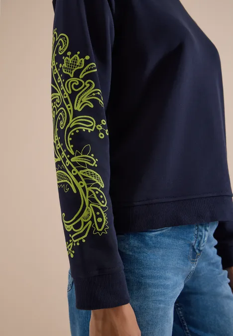 Damenpullover in dunkelblau mit neon-grünem Paisley-Muster auf dem Ärmel, kombiniert mit Jeans