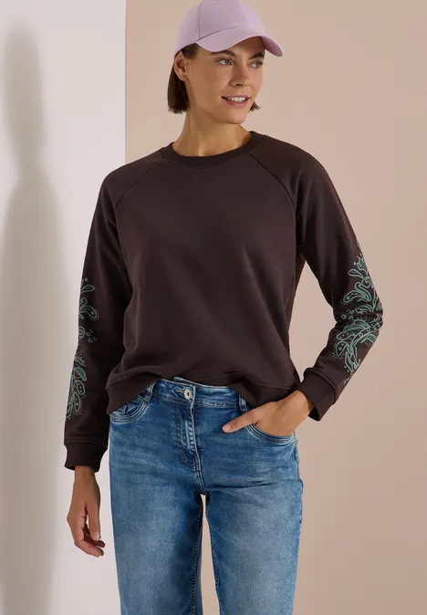 Frau in lässigem Outfit: dunkles Sweatshirt mit Musterärmeln, Jeans und lila Cap, lächelnd vor neutralem Hintergrund.