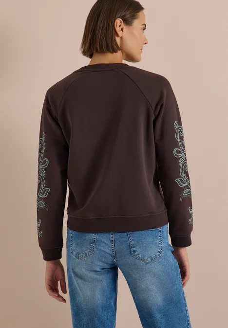 Frau von hinten in dunkelbraunem Sweatshirt mit türkisfarbener Blumenstickerei an Ärmeln und blauen Jeans.