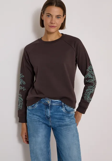 Frau in Jeans und braunem Langarmshirt mit grünen Mustern an den Ärmeln vor neutralem Hintergrund.