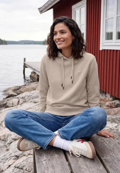 Sweatshirt met lange mouwen en capuchon snow cloud beige Frau sitzt entspannt im Schneidersitz auf Holzsteg vor rotem Haus am See.