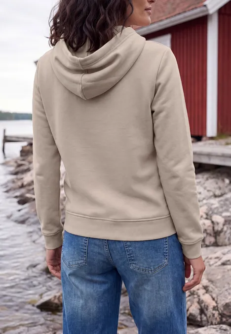 Sweatshirt met lange mouwen en capuchon snow cloud beige Person in beigefarbenem Hoodie und Jeans steht am felsigen Ufer vor einem roten Bootshaus.
