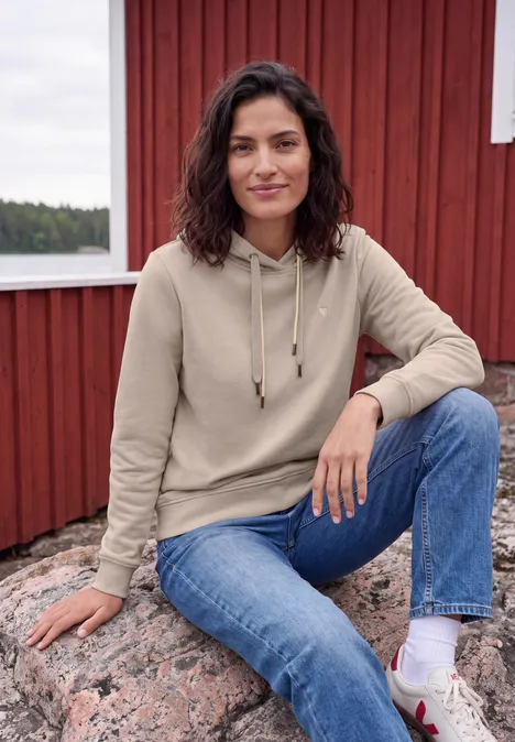 Sweatshirt met lange mouwen en capuchon snow cloud beige Eine Frau in beiger Kapuzenjacke und Jeans sitzt auf einem Felsen vor roter Holzwand und lächelt.