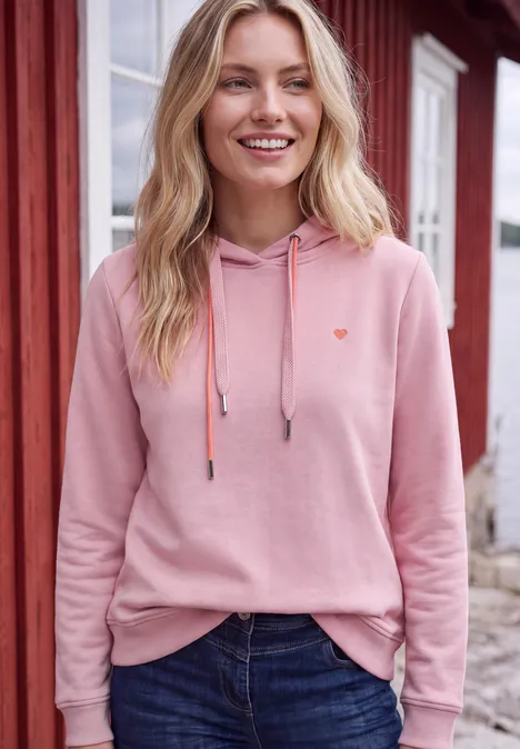 Sweatshirt met lange mouwen en capuchon mellow rose Lächelnde Frau in rosa Kapuzenpullover und Jeans steht vor einem roten Haus.