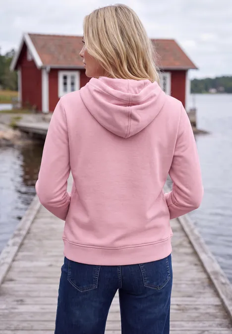 Sweatshirt met lange mouwen en capuchon mellow rose Frau mit rosa Hoodie und Jeans steht mit dem Rücken zur Kamera auf einem Steg am See vor einem roten Haus.