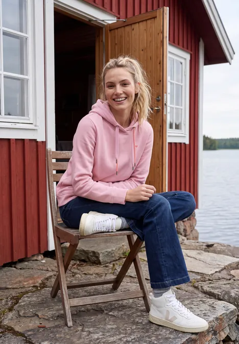 Sweatshirt met lange mouwen en capuchon mellow rose Frau in rosa Kapuzenpullover sitzt auf Holzstuhl vor roter Hütte am See, lächelnd und entspannt.