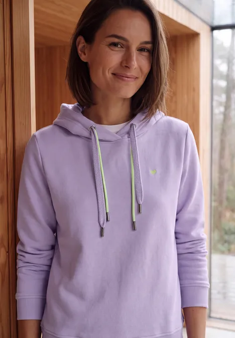 Langarm Sweatshirt mit Kapuze cool lilac Frau mit kurz geschnittenem Haar und lila Kapuzenpullover, die lächelnd vor einer Glasfront steht.